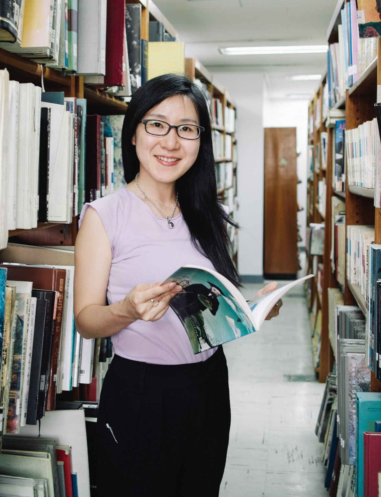Woman in glasses holding a magazine in a library aisle lined with books.