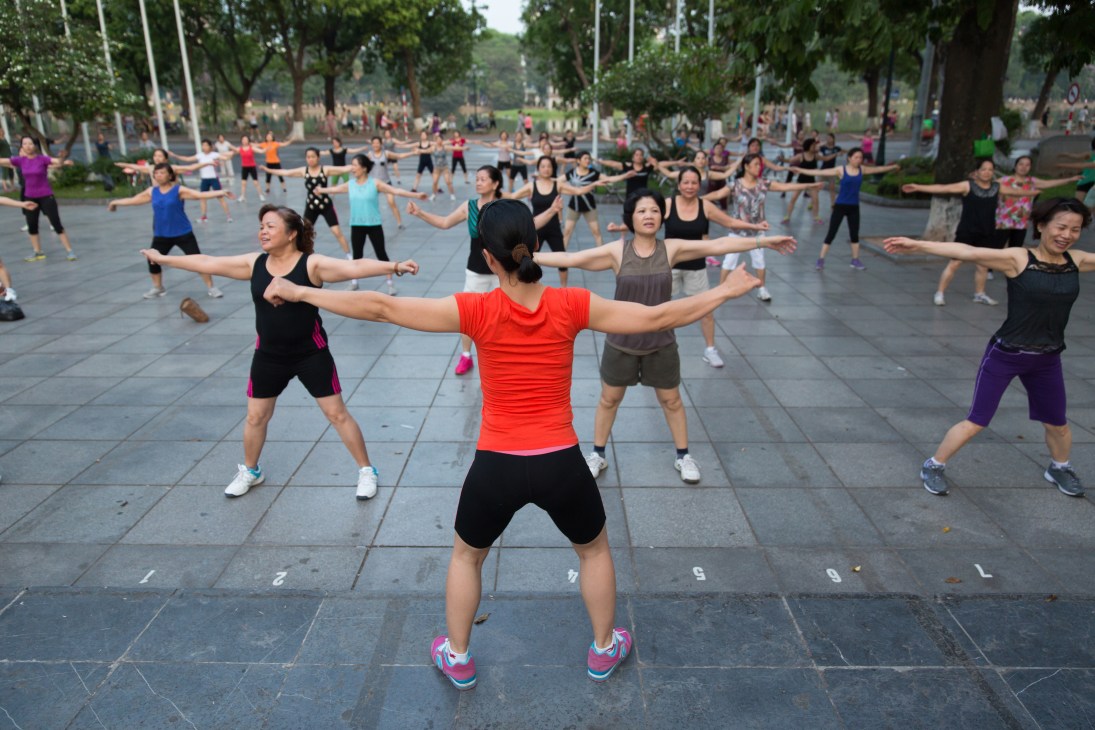 Group fitness class exercising in Hanoi's Old Quarter with large trees and colonial buildings in background.