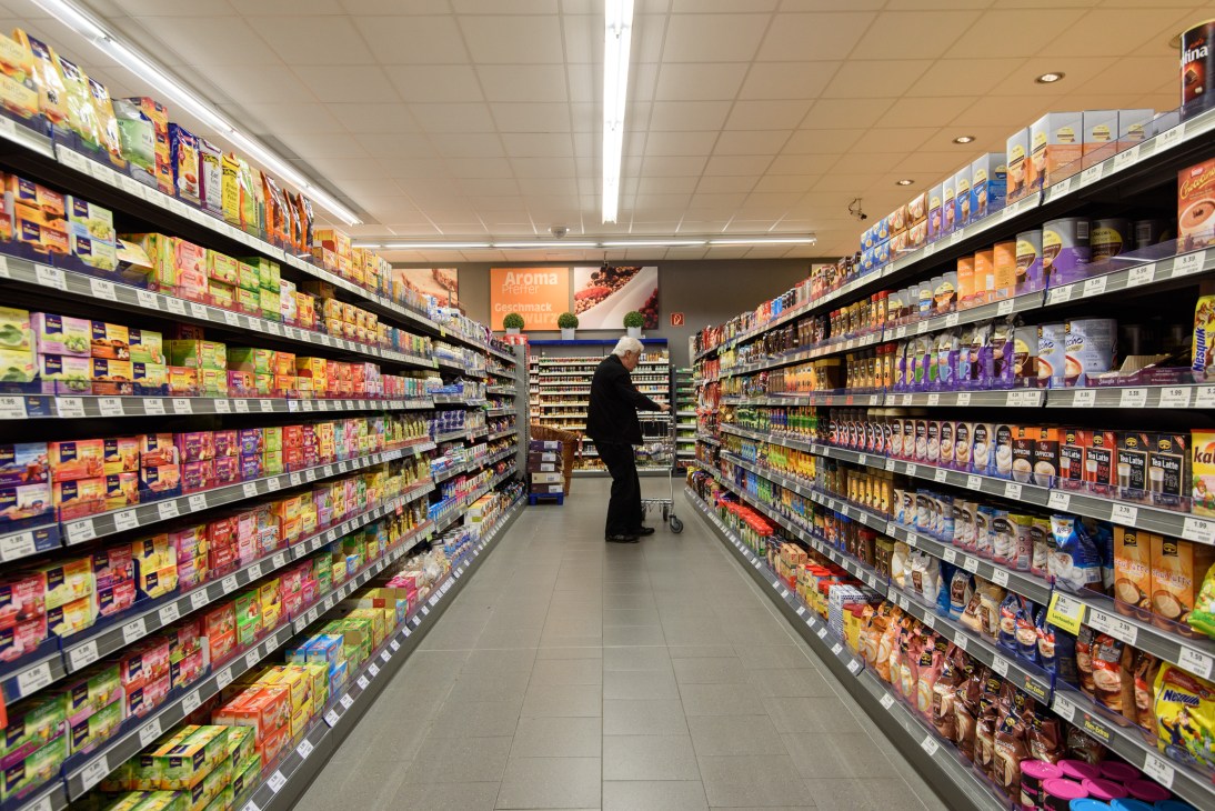 Interior of a German supermarket with fully stocked shelves of colorful packaged products lining both sides of an aisle, with a shopper visible in the distance.