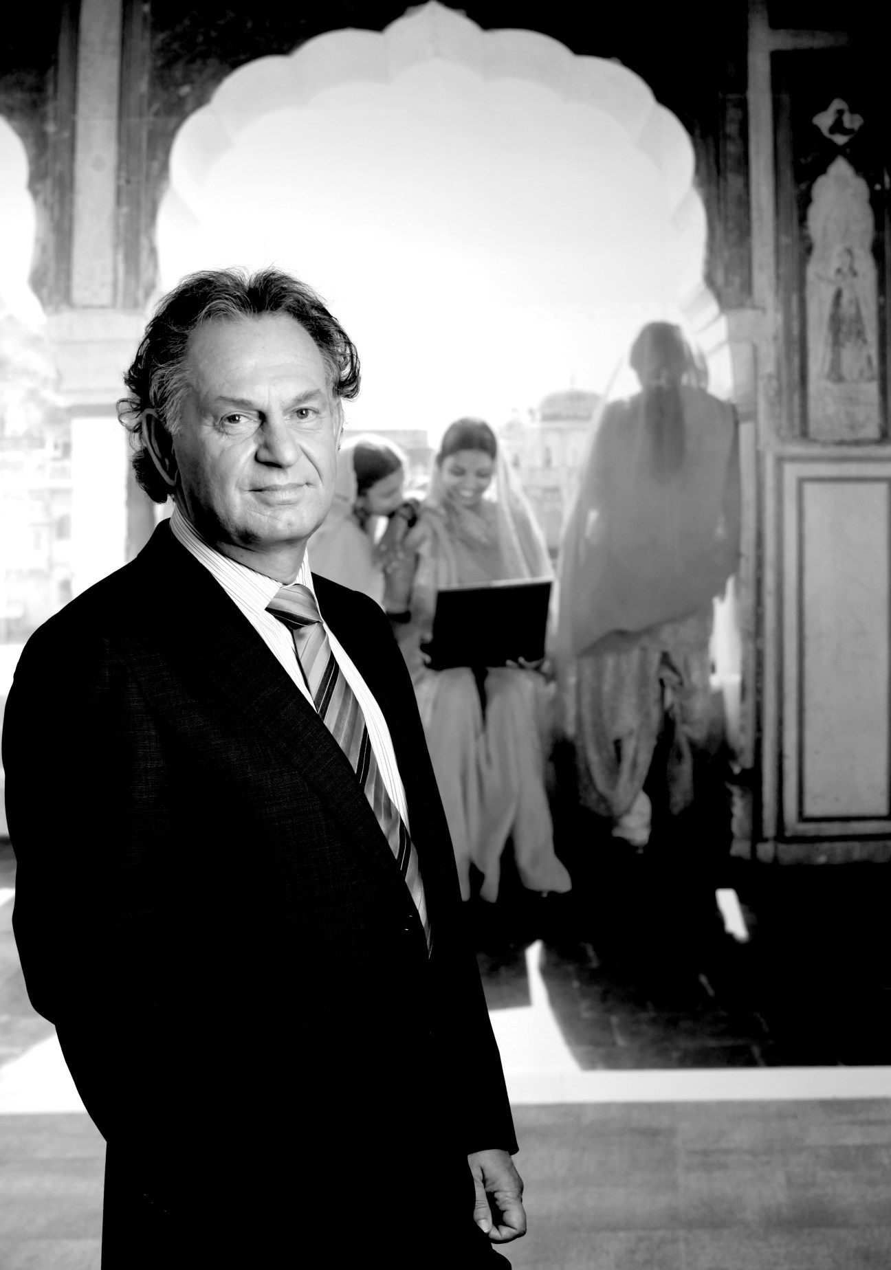 Black and white portrait of a man in business suit and striped tie standing in an archway with blurred figures in background.