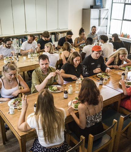 Staff members gathered around wooden tables eating lunch together in a bright office space with shelving in the background.