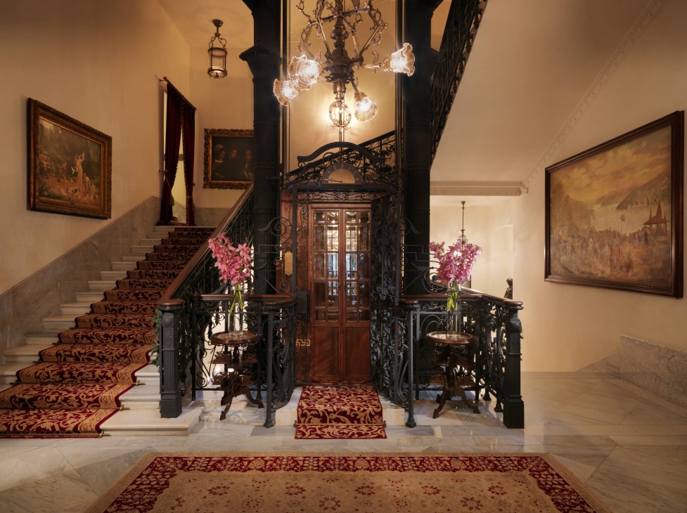 Grand staircase with ornate black iron railings, patterned carpet runner, crystal chandeliers, and framed paintings on pale walls in a historic hotel lobby.
