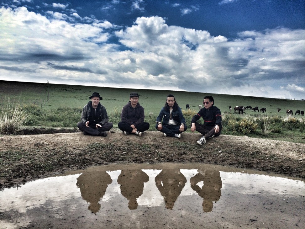 Four band members sitting on ground with their reflections in muddy water, rolling hills and cattle in background.