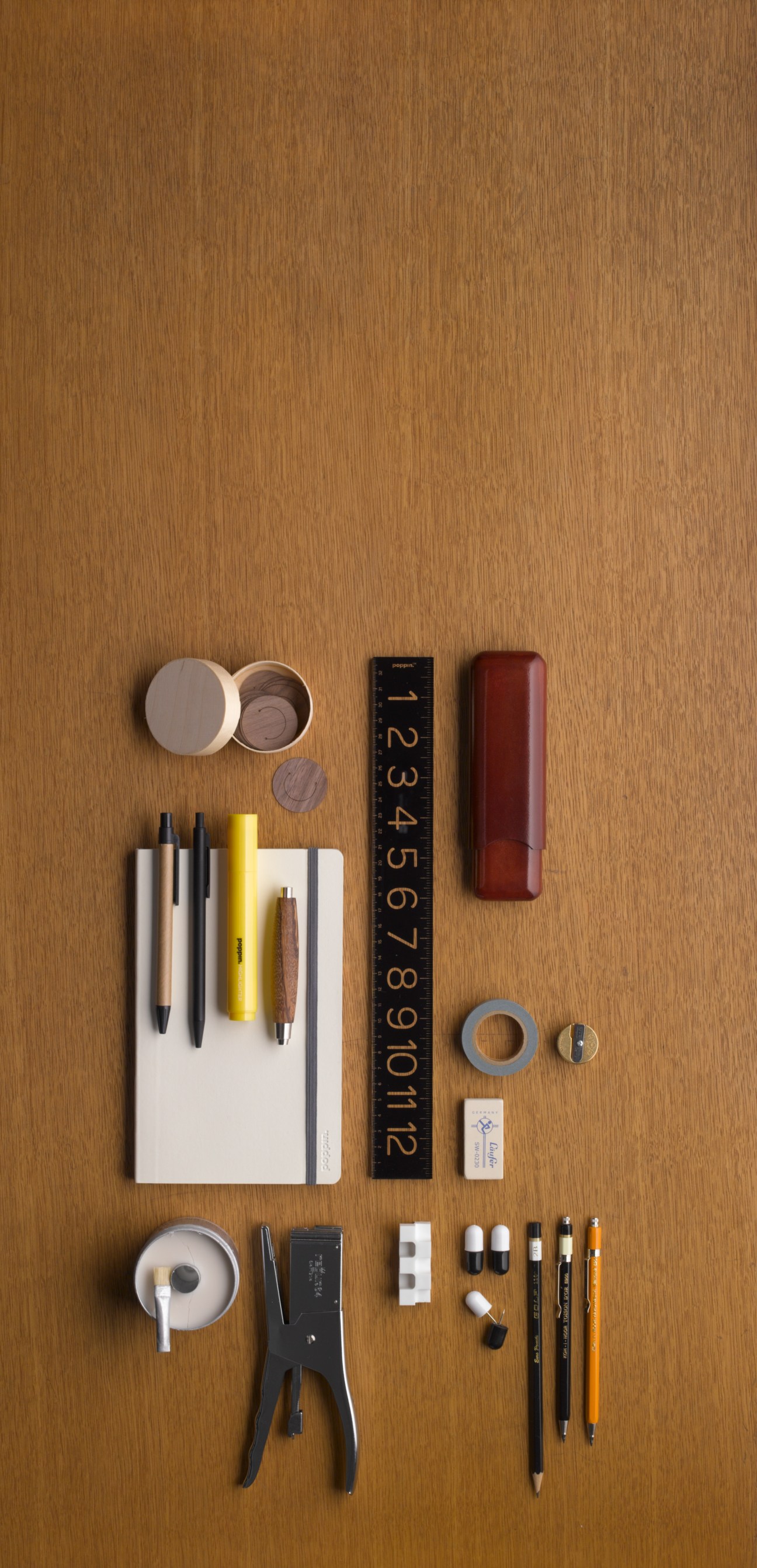 Flat lay of stationery items including pens, ruler, eraser, notebook, tape, and leather case on wooden surface.