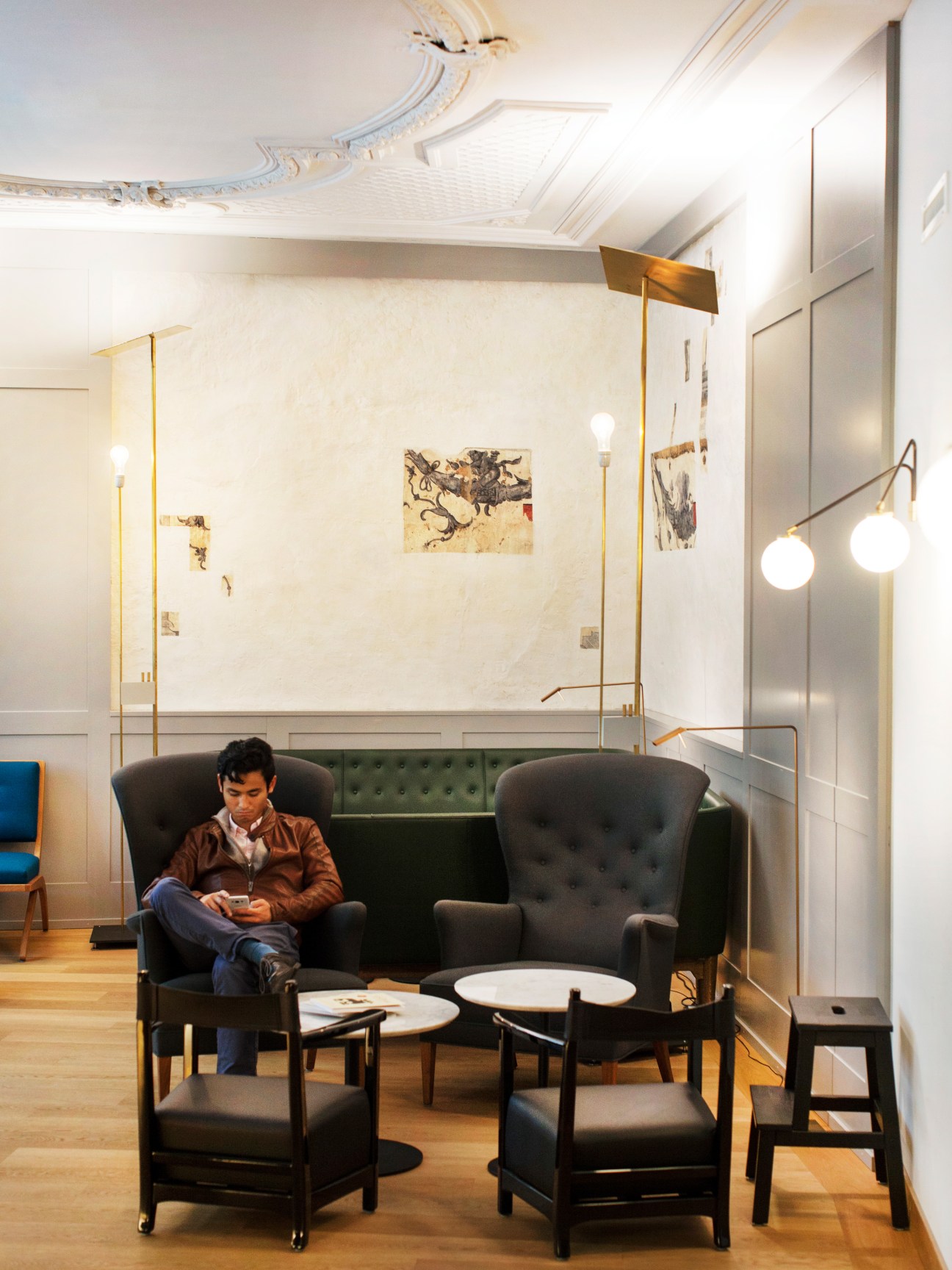 Modern hotel lounge with a man seated in a large black wingback chair next to a green sofa, brass floor lamps, and white walls with artwork.
