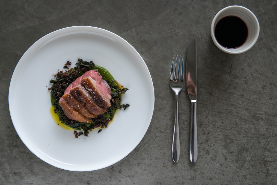Roasted lamb rump with nettle sauce and dried olives plated on white dish with fork and knife.