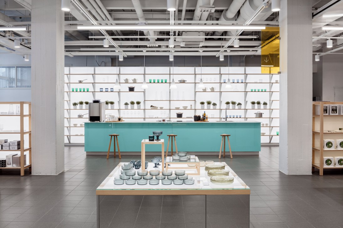 Modern retail showroom with turquoise counter, white shelving displaying ceramics and kitchenware, and wooden display tables.