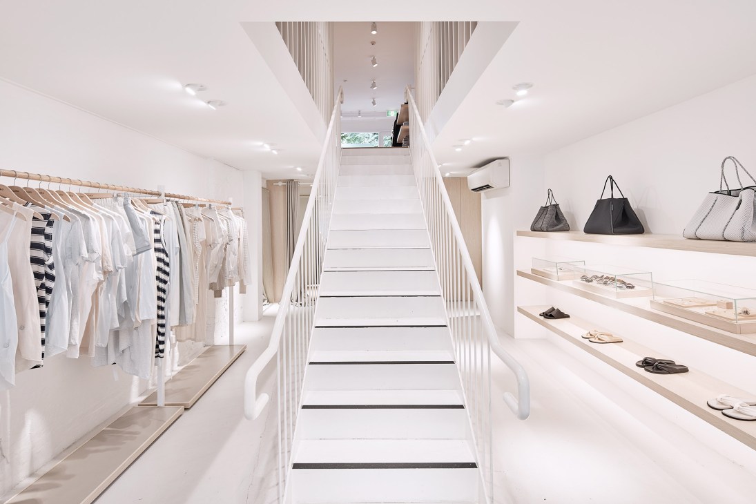 Minimalist retail interior with white walls, wooden clothing racks on left, central staircase with metal railings, and display shelves with handbags and shoes on right.