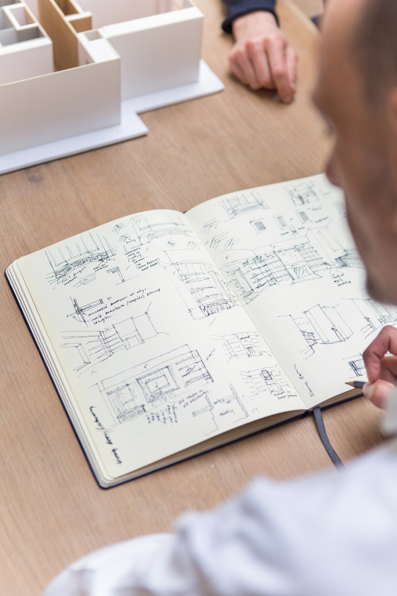 Architect's hand pointing to sketches in an open notebook with architectural drawings and annotations.