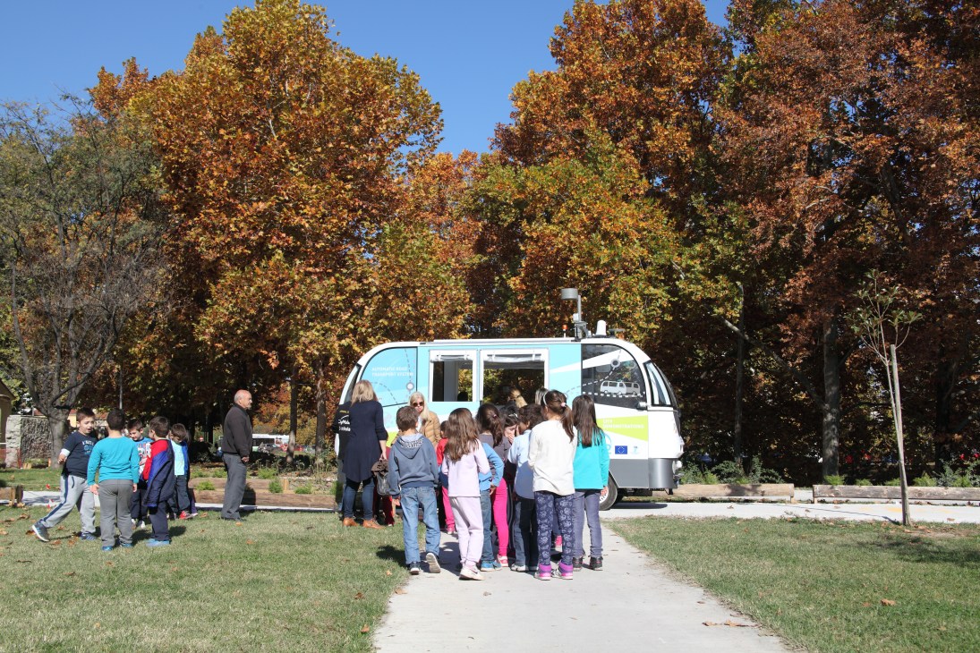 A white and light blue autonomous shuttle bus with people boarding at a park with autumn trees.