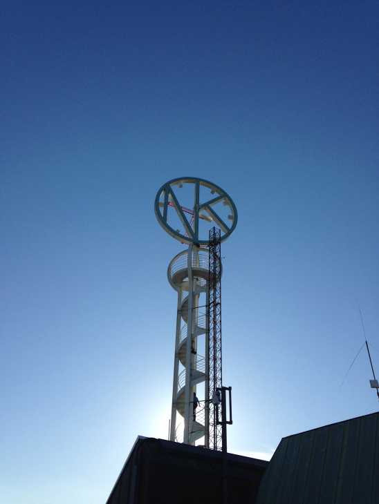 A tall architectural clock tower structure against a blue sky with a large circular clock face at the top.