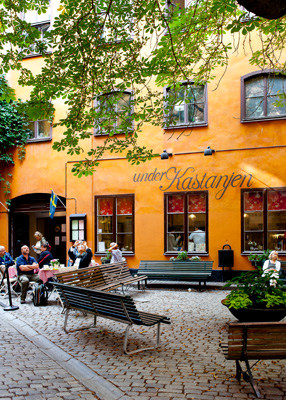 Orange building facade with ivy vines, outdoor seating area with benches and people gathered in a European courtyard
