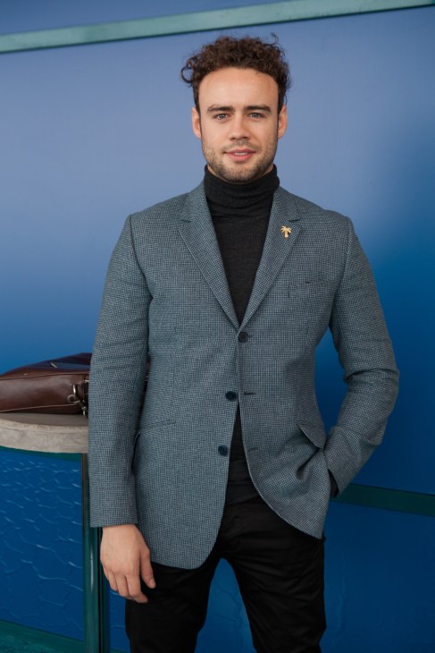 Man in grey blazer and black turtleneck standing against blue background.