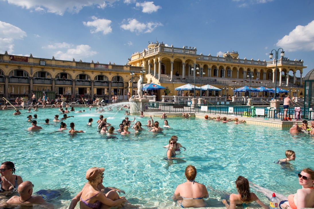 Swimmers enjoying the turquoise outdoor pool at Széchenyi baths in Budapest with ornate yellow buildings and blue umbrellas.