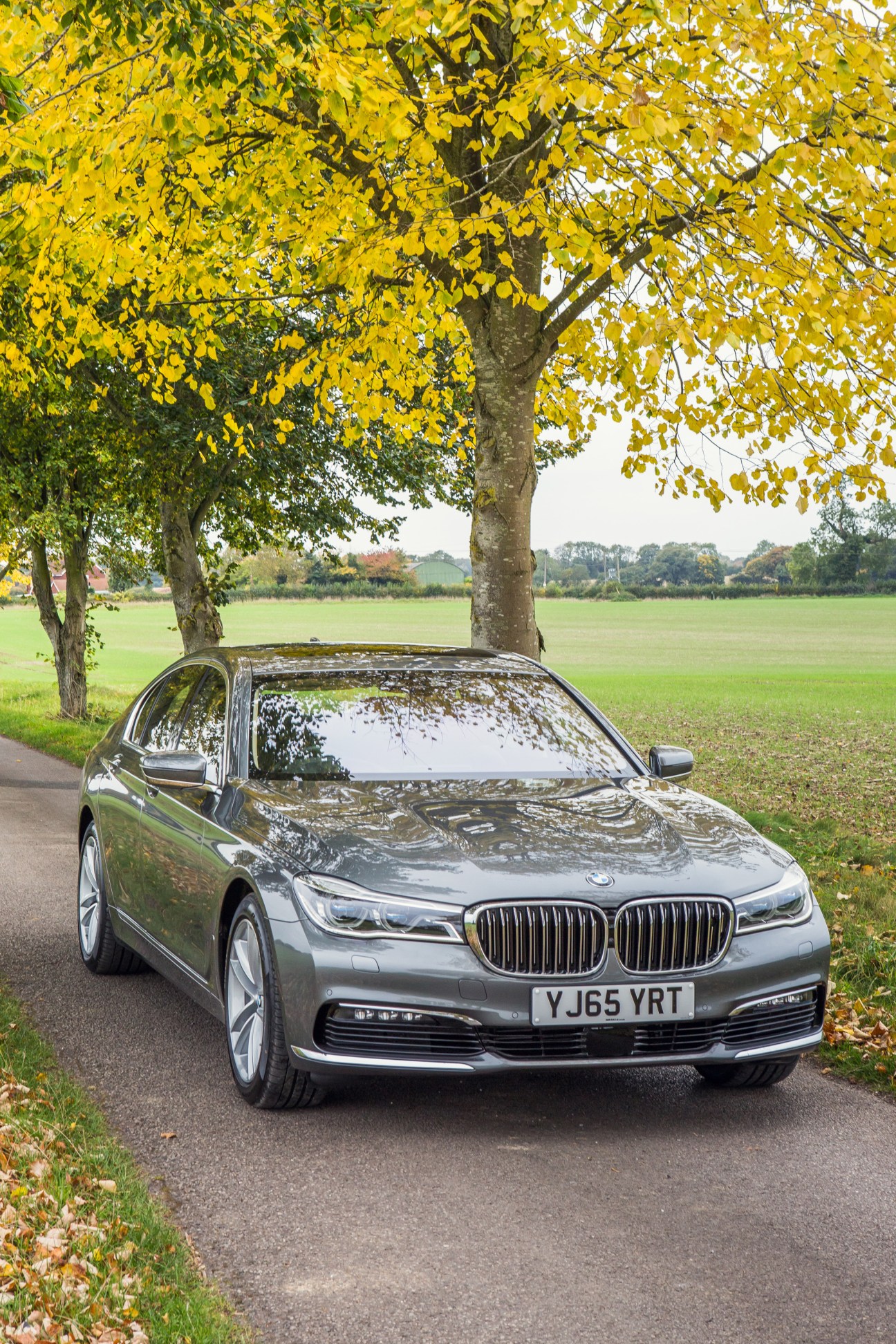 Silver BMW 7 Series parked on a tree-lined rural road with golden autumn foliage overhead.