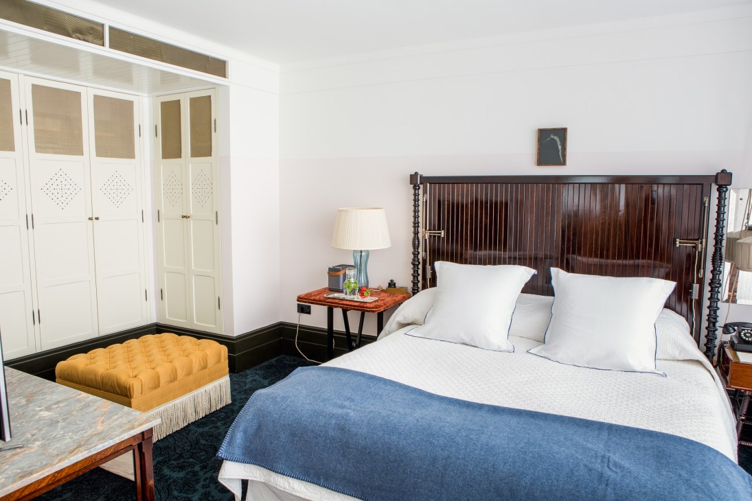 Luxurious bedroom with dark wooden bed frame, white linens, yellow ottoman, and cream-colored wardrobe at Chiltern Firehouse, London.