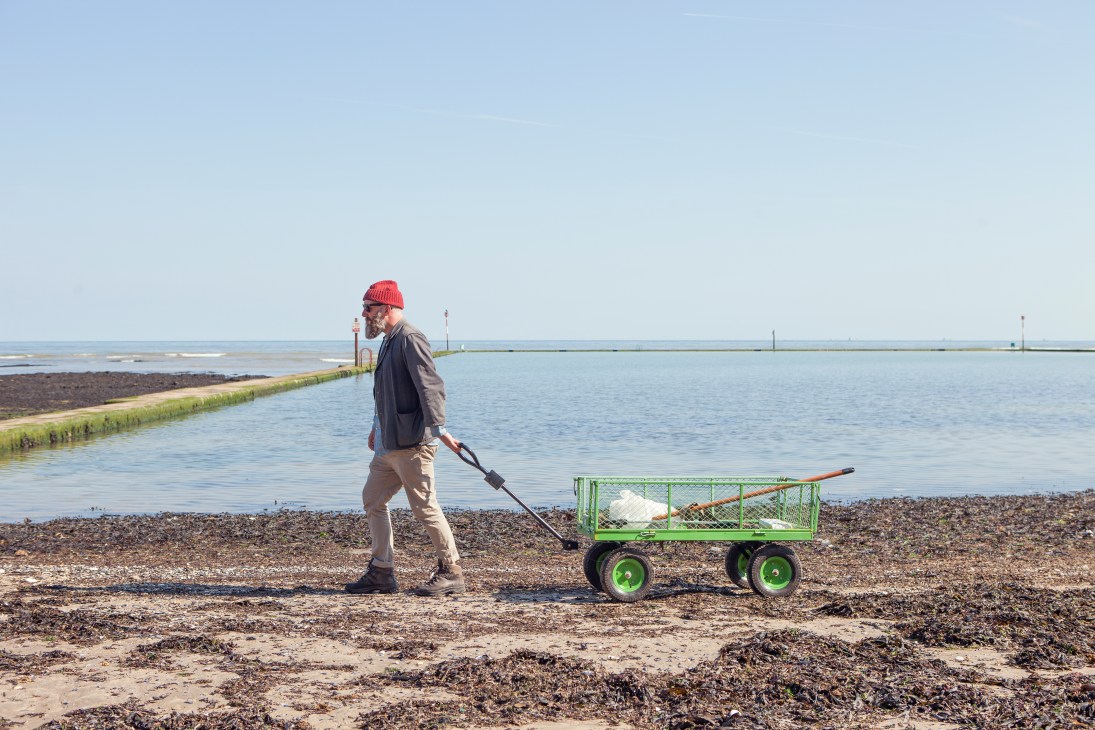 Man in red beanie harvesting seaweed on a beach with a green cart and rake at low tide.