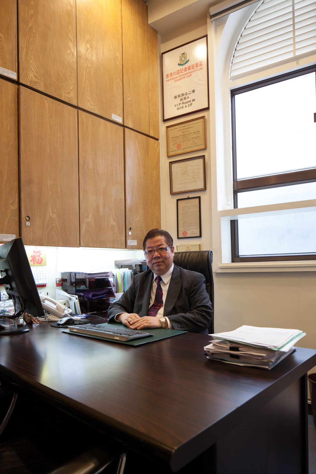 Man in business attire seated at desk in wood-paneled office with framed certificates on wall
