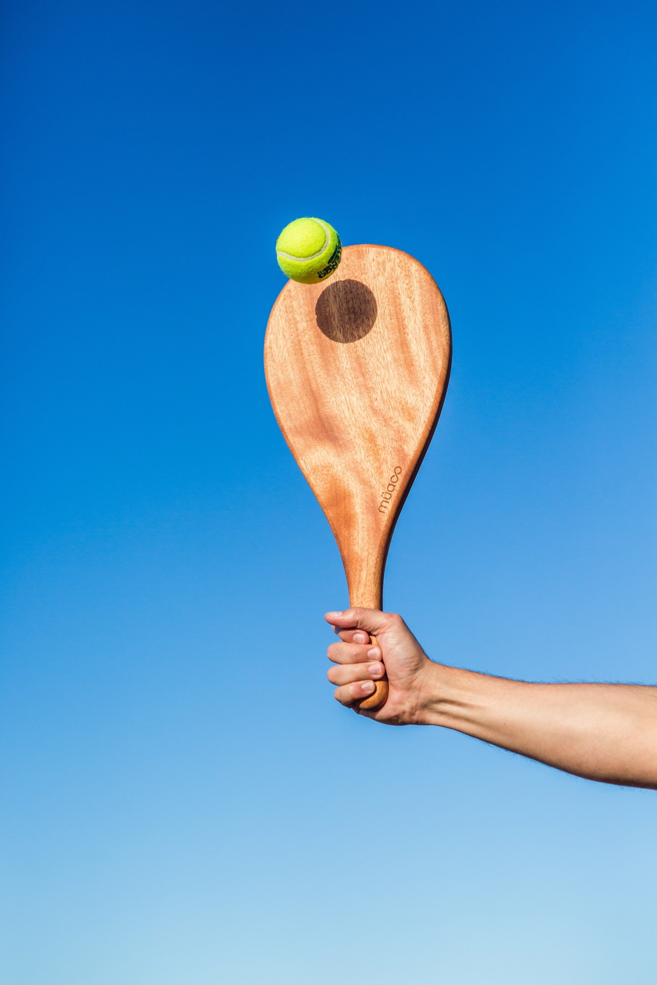 Wooden pádel bat with yellow ball against bright blue sky held in hand