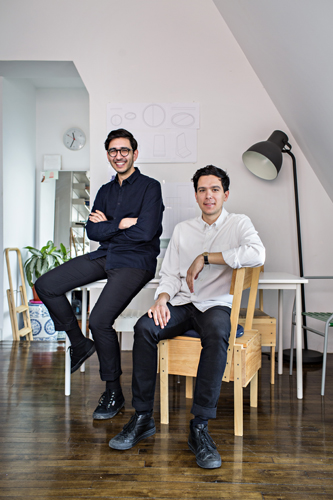 Two men pose in a bright studio space with wooden chairs, a potted plant, and a black floor lamp.
