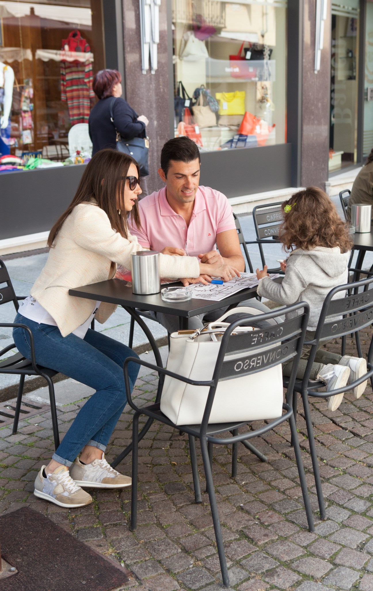 A family sits at an outdoor café table on a pedestrian street, with storefronts visible in the background.