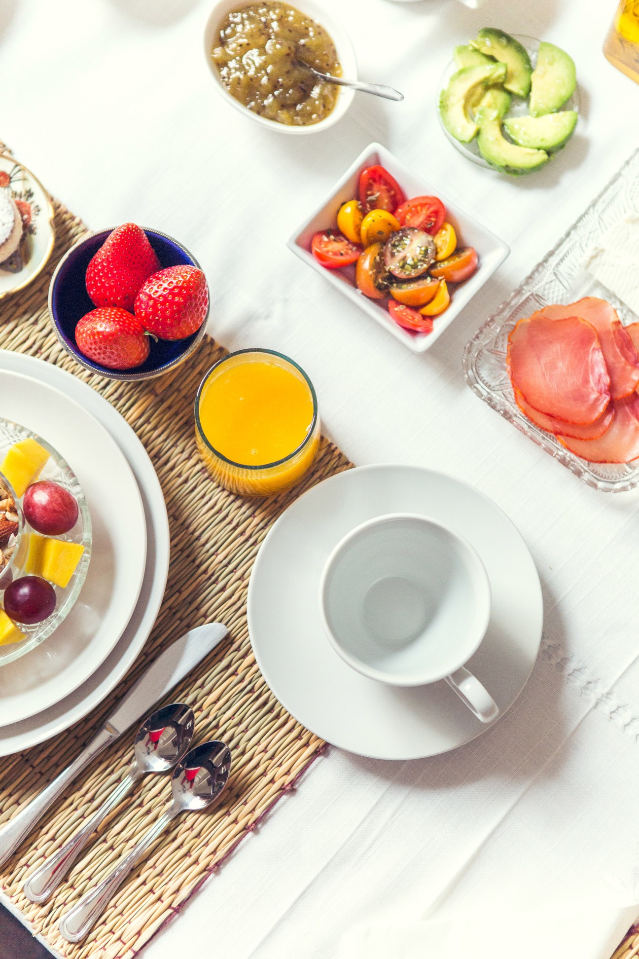 Overhead view of a colorful breakfast spread with strawberries, sliced tomatoes, cucumber, cured meat, juice, and pastries on white dishes