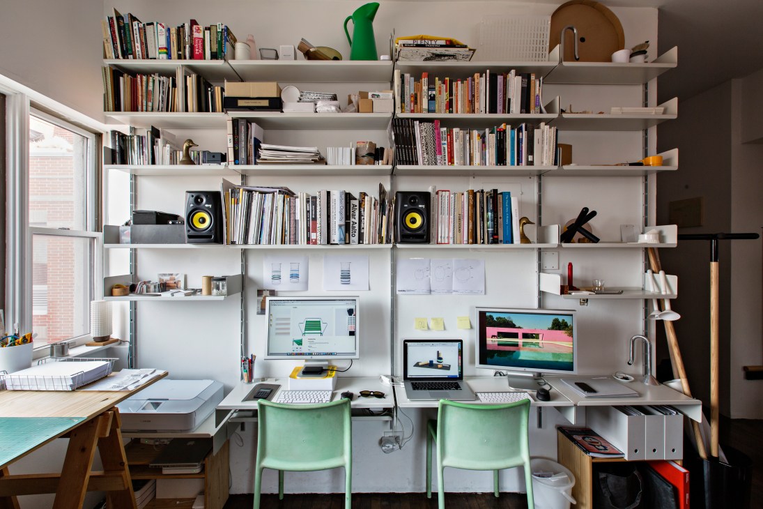 A modern home office workspace with white shelving, green chairs, and dual monitors displaying design work.