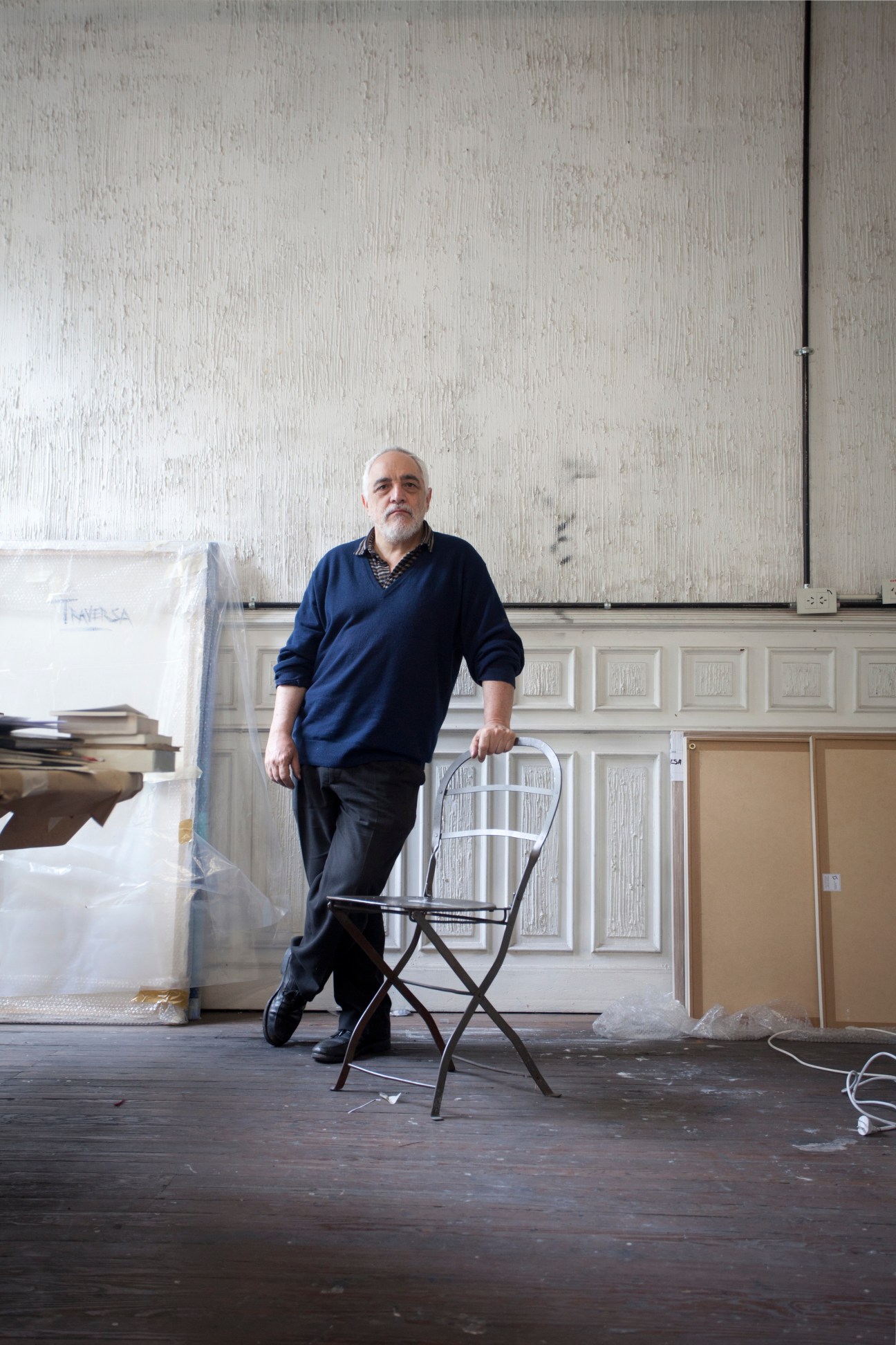 Man standing in an artist's studio holding a chair, with textured white walls and wooden floors.