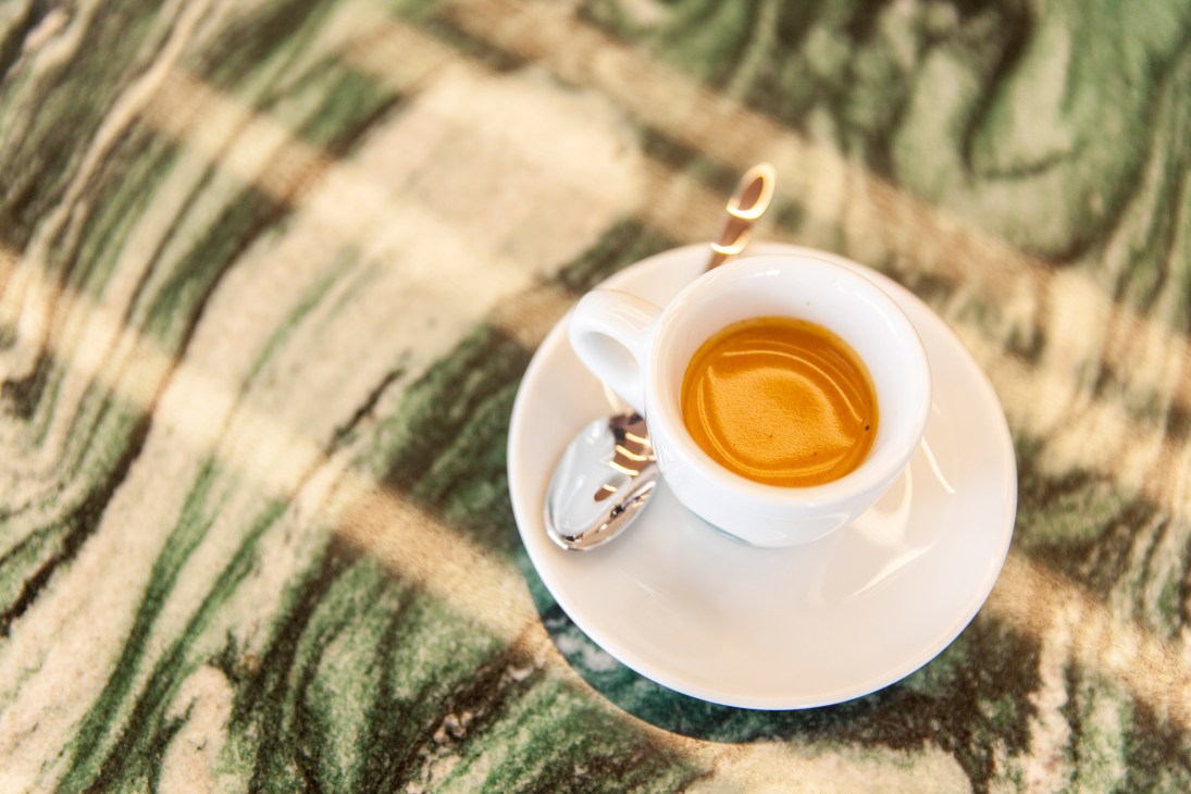 Overhead view of a white cup of espresso with latte art on a saucer with spoon on a striped green and beige table.