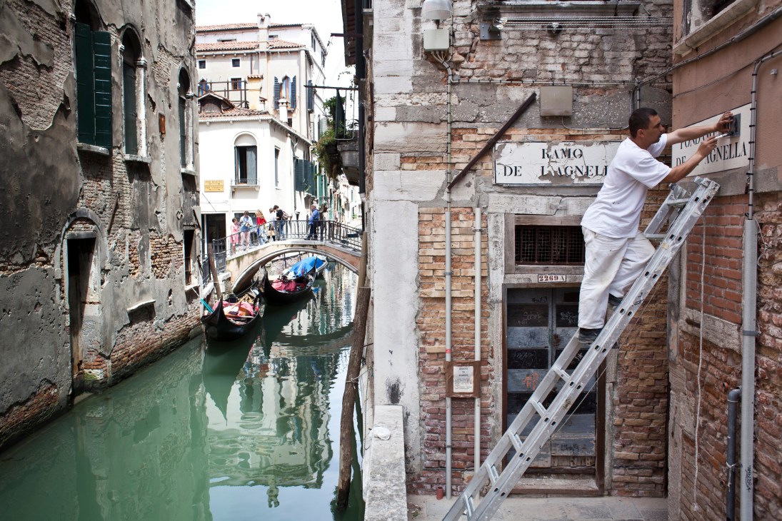 Worker on ladder restoring a whitewashed nizioleti street sign in Venice with canal and gondolas below.