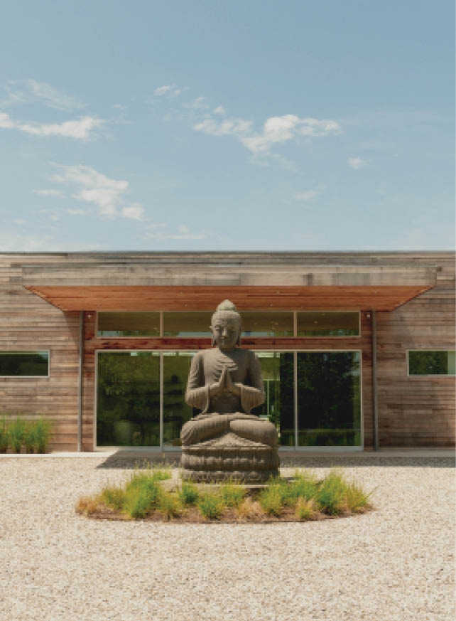 Buddha statue in pebble garden before wooden-shingled wellness retreat building with large windows.
