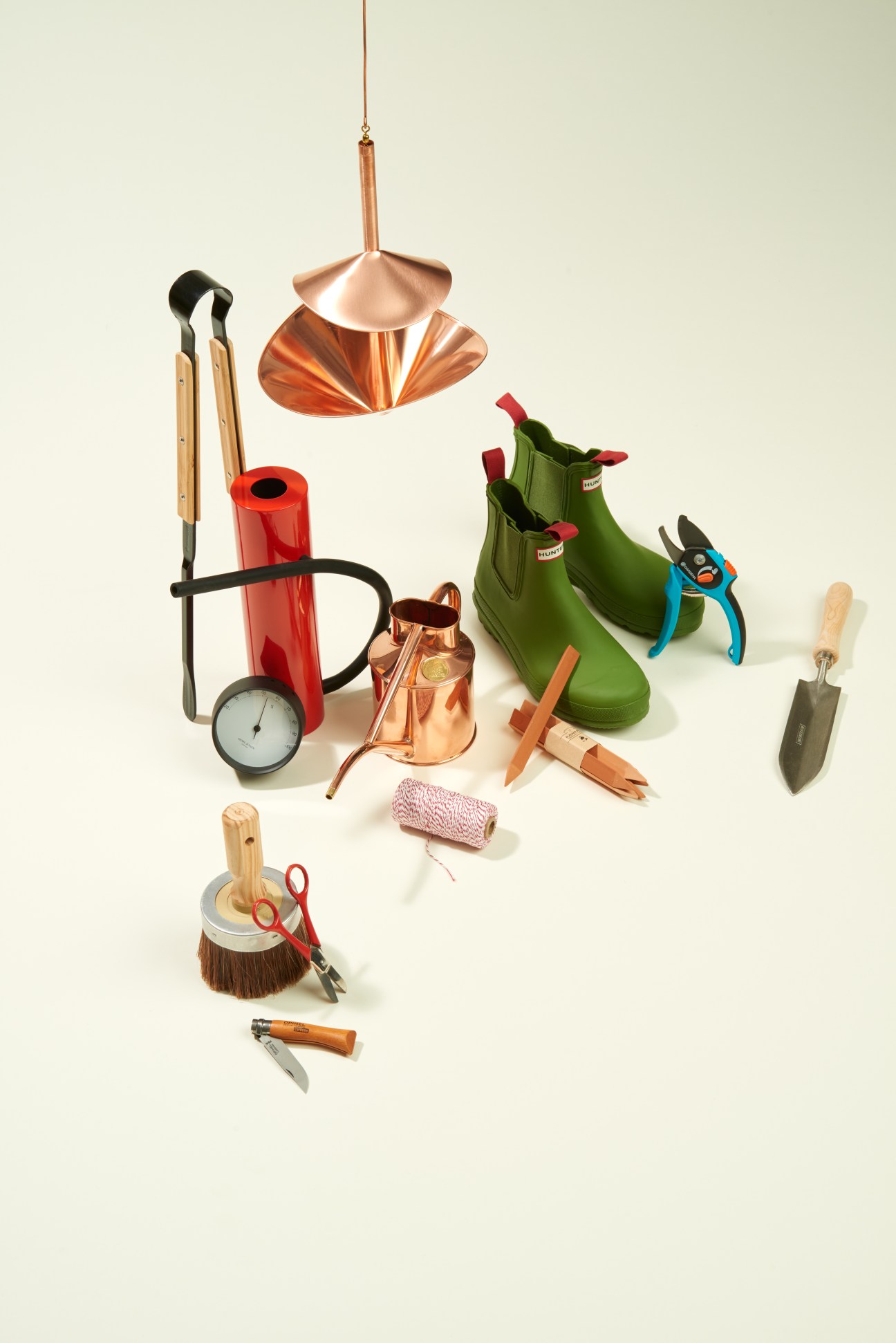 Curated garden tools and accessories including green wellington boots, copper watering can, secateurs, trowel, and twine arranged on a white background.