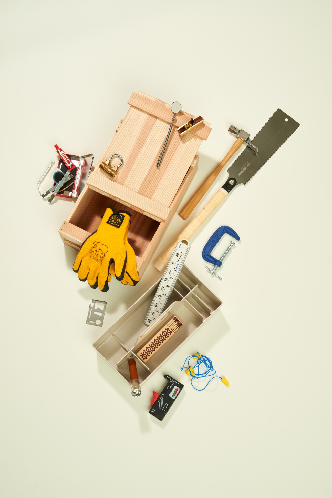 Collection of hand tools including wooden toolbox, yellow work gloves, hammer, saw, ruler, clamp, and measuring devices arranged on beige background.