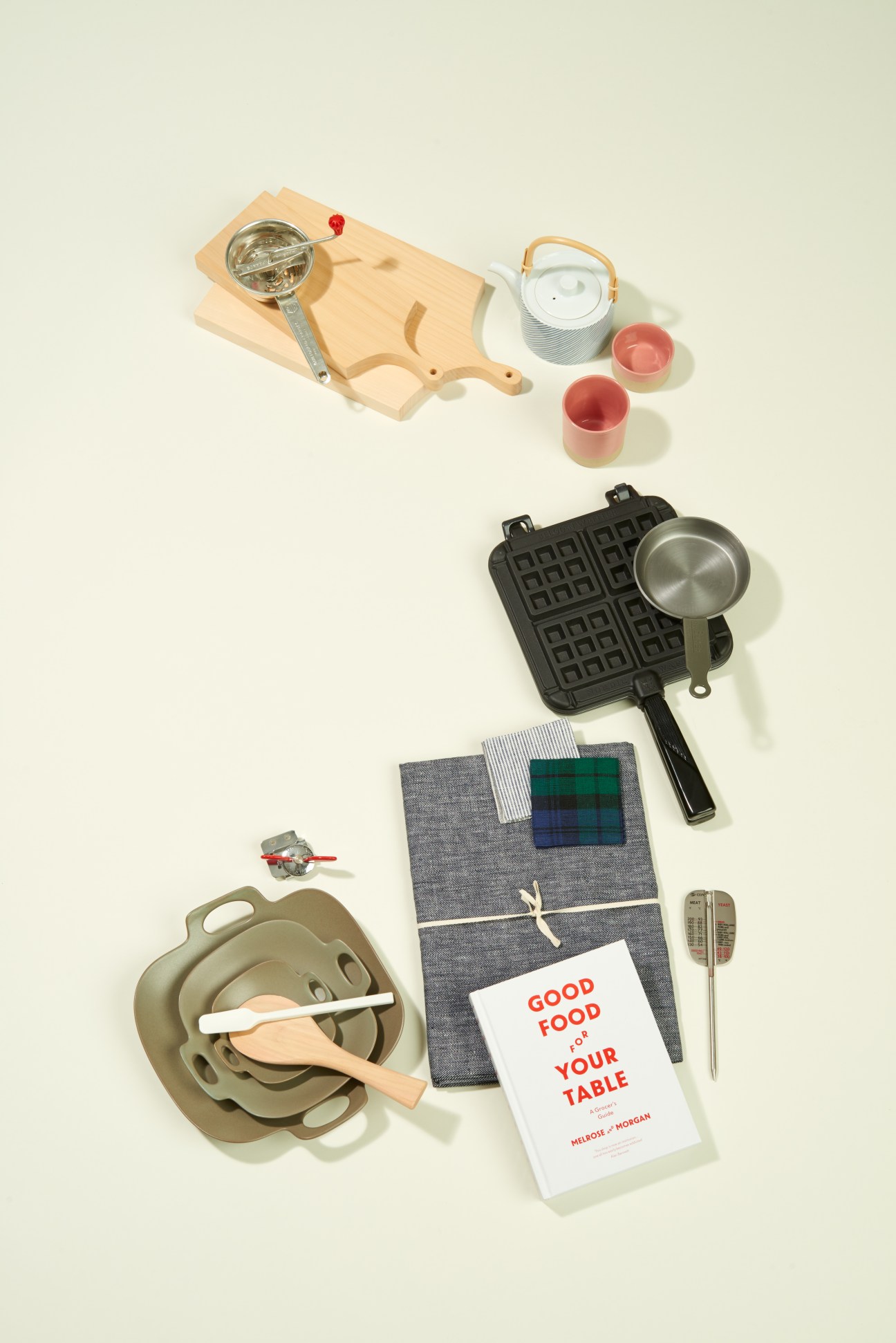 Kitchen items arranged flat lay including wooden chopping board, waffle maker, teapot, pink cups, tablecloth, and cookbook.