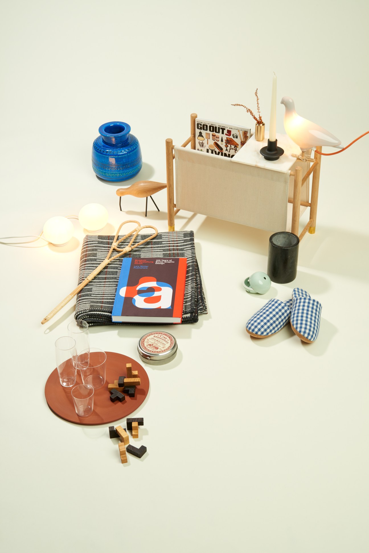 Styled arrangement of home goods including a wooden organiser, ceramic vase, pigeon light, gingham slippers, and various decorative items.