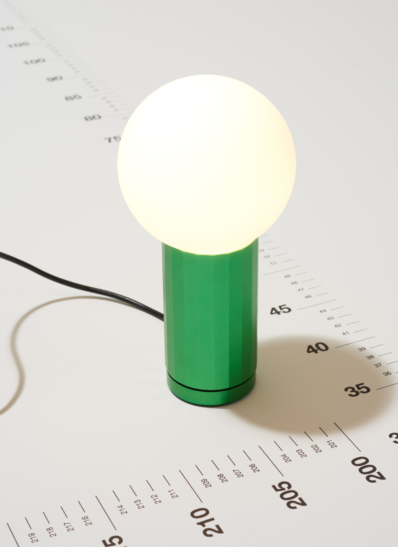 Turn On desk lamp by Wrong for Hay with green cylindrical base and white spherical light head on ruler.
