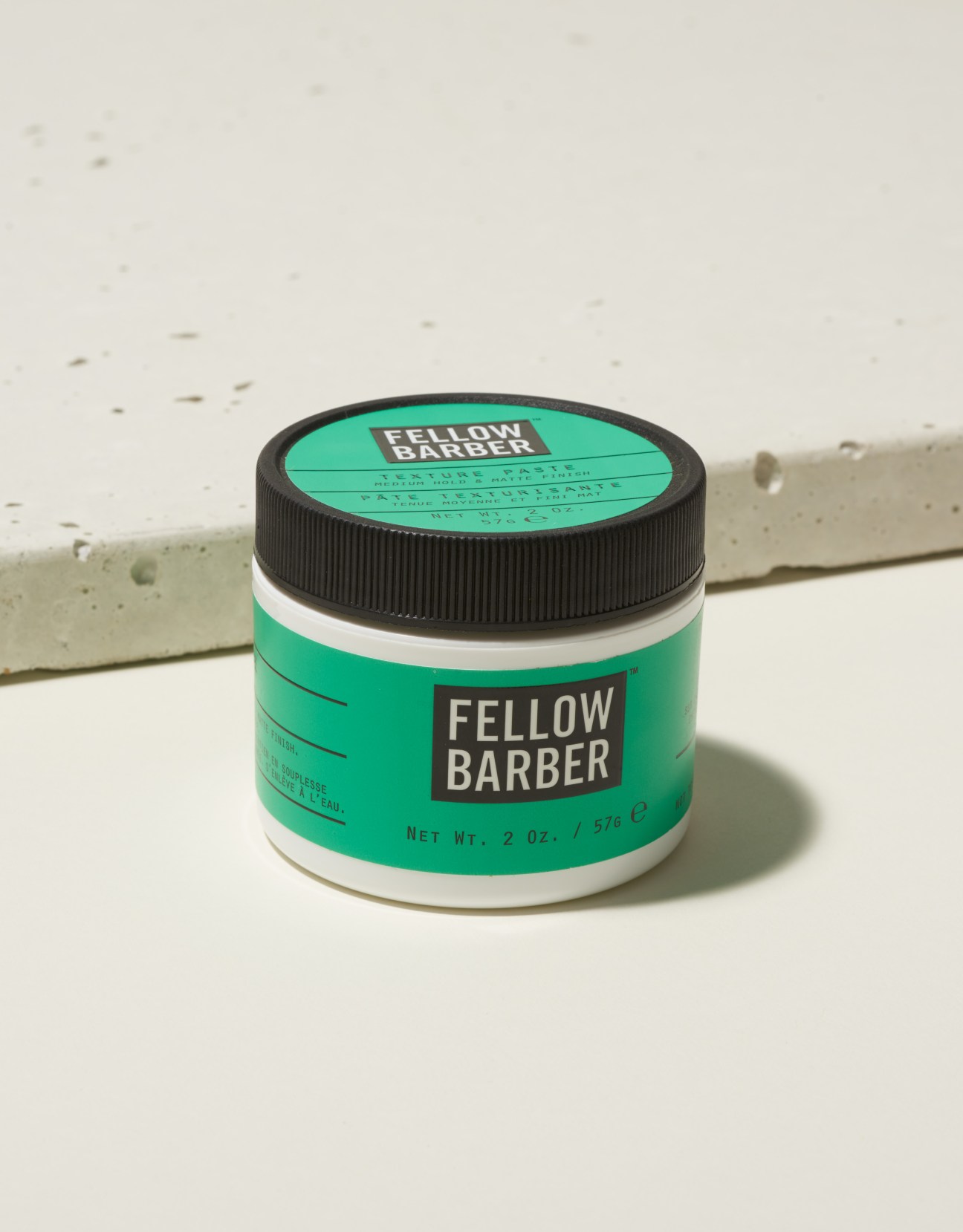 Fellow Barber hair wax in green packaging with black screw-top lid on concrete surface.