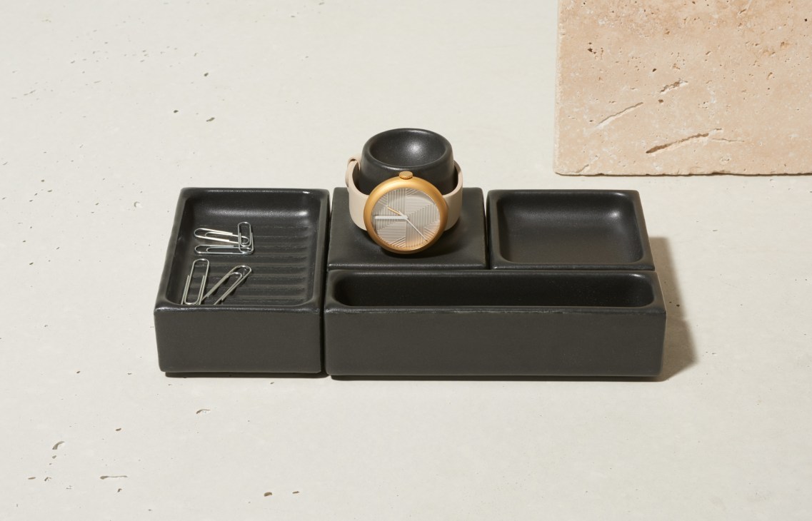 Objest watch with copper bezel and numberless face displayed on black porcelain desk organizer with paper clips.