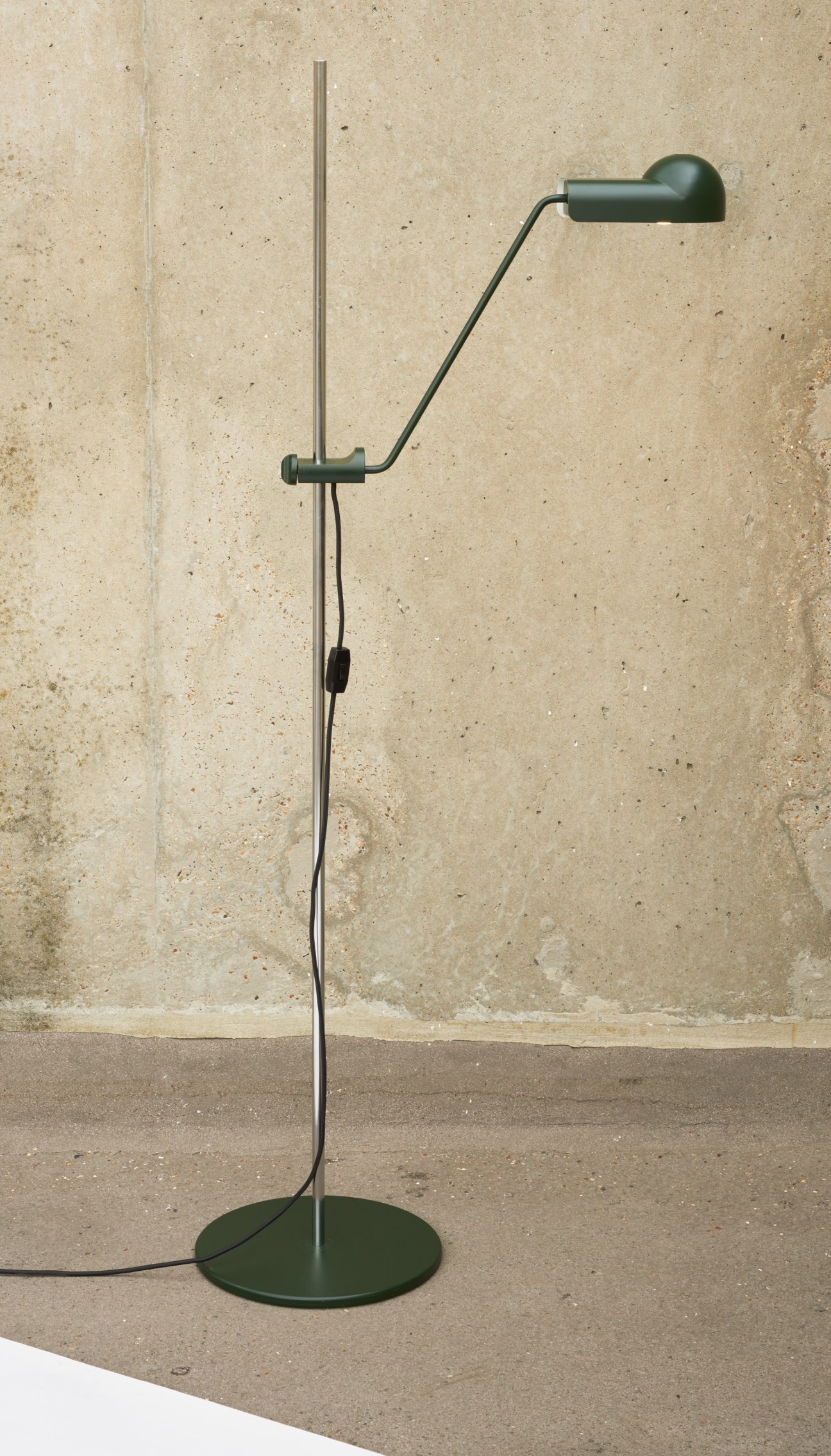 Green angular floor lamp with adjustable head mounted on tall stand against textured wall