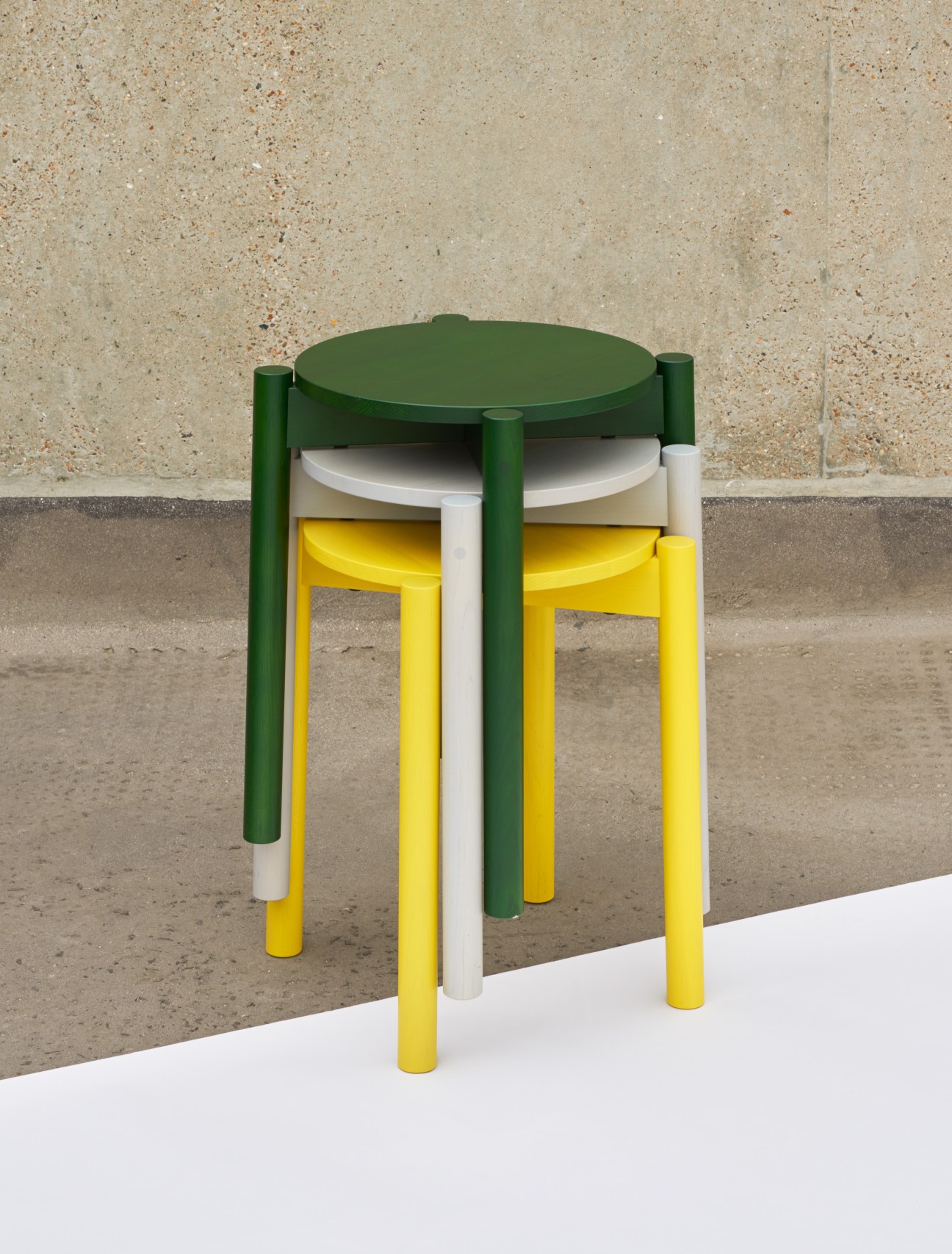 Four colorful Castor Stool Plus stools stacked together in green, white, and yellow against concrete wall