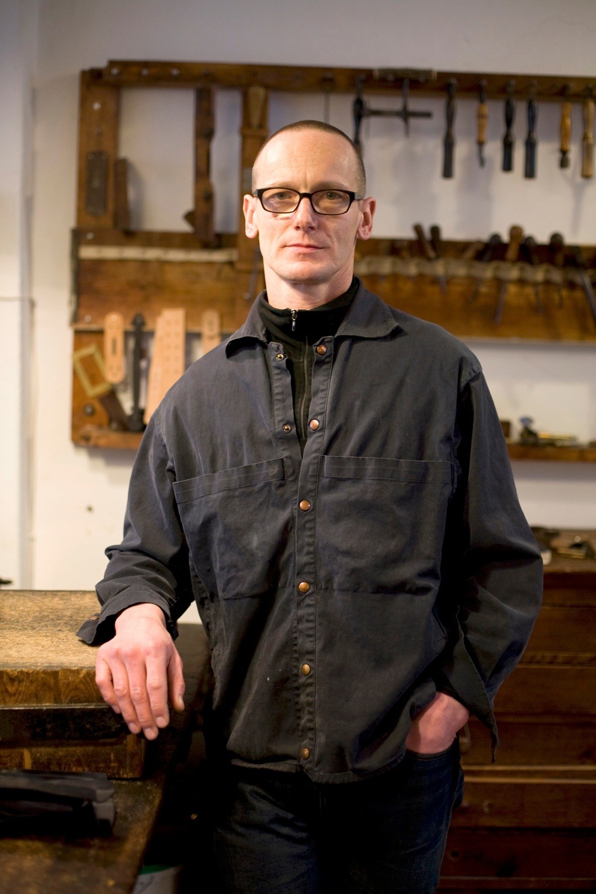 Jörn Rischke, leather goods designer and manufacturer, stands in his workshop with tools and workbenches visible behind him.