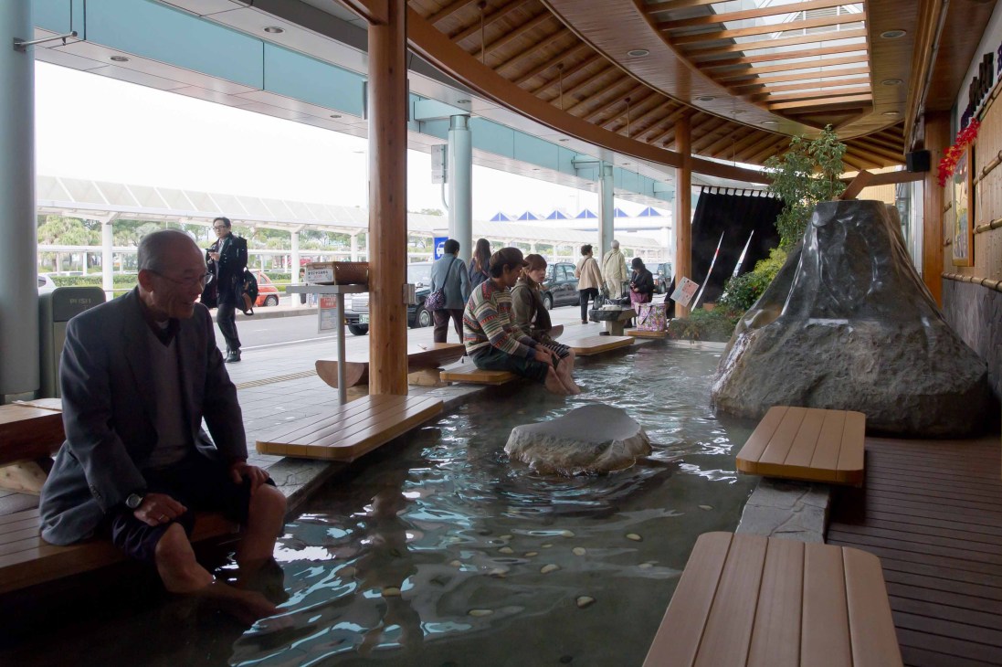 Visitors enjoying a hot spring foot bath at Kagoshima Airport's departures area with wooden seating and natural rock features.