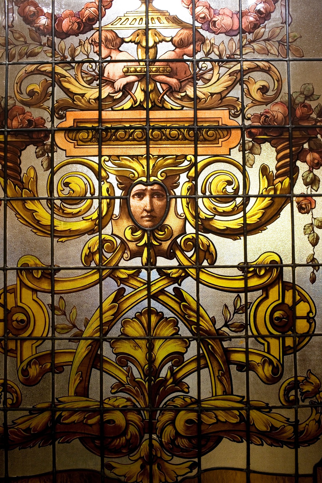 Ornate stained glass window with golden scrollwork, roses, and a classical face motif in the center.