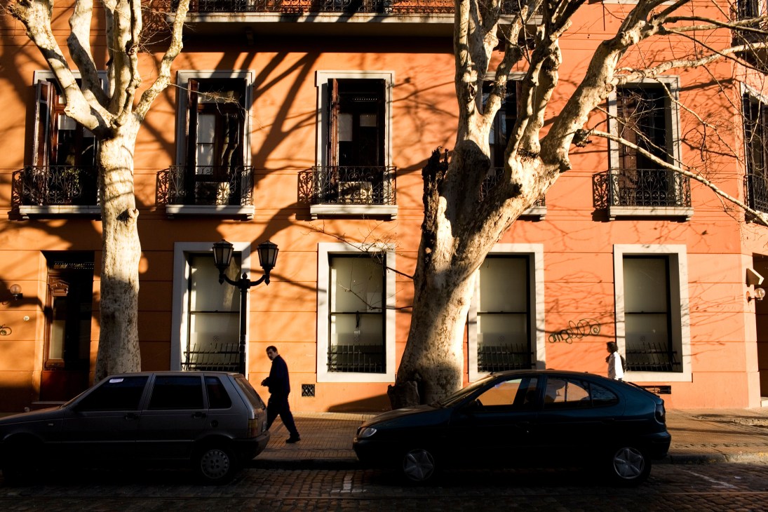 Orange colonial building in Montserrat with wrought-iron balconies, large trees, and parked cars on street.
