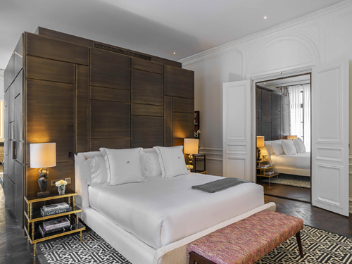 Hotel bedroom with dark wood headboard, white bedding, geometric patterned rug and orange bench
