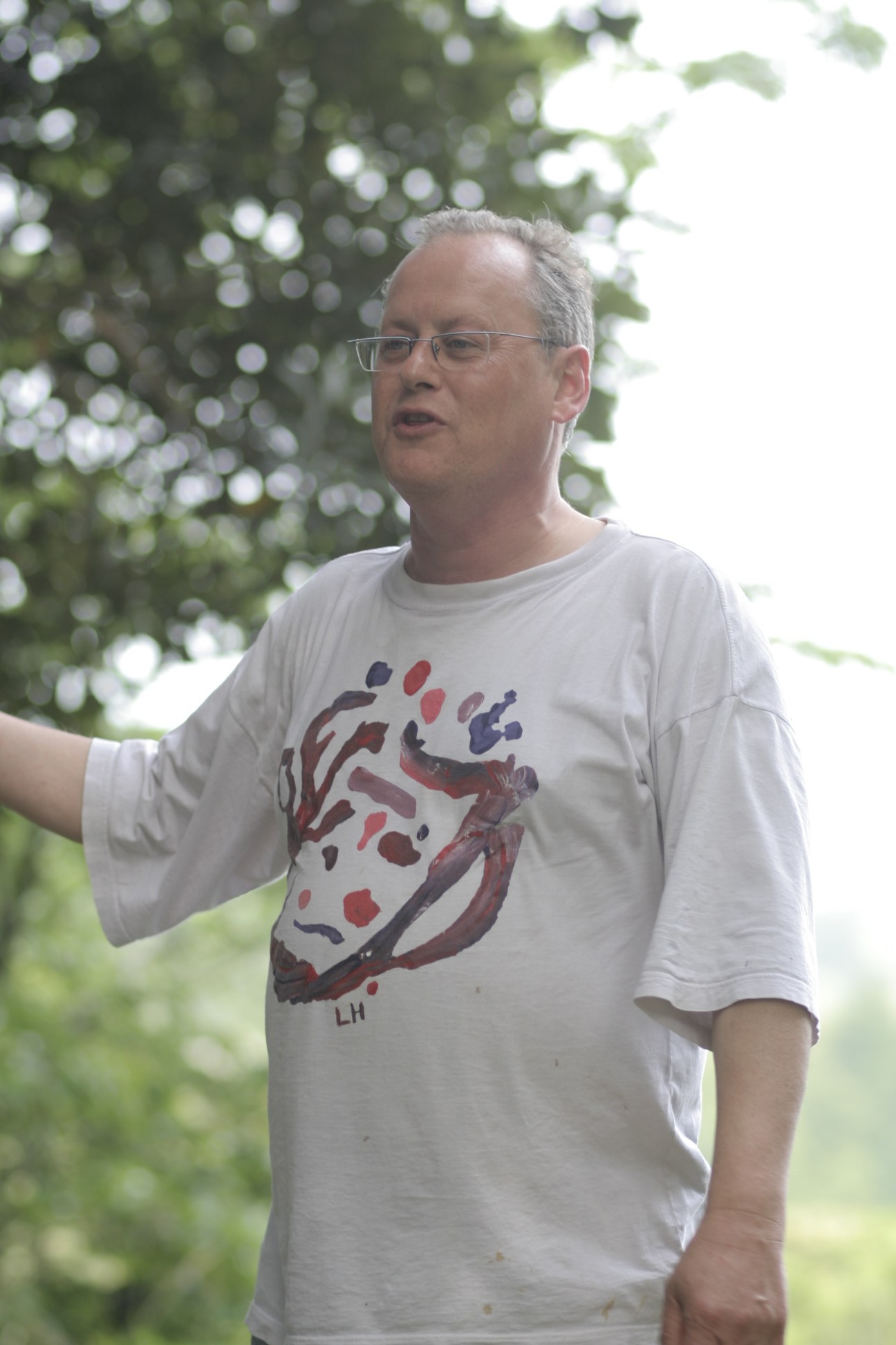 Man wearing glasses and graphic t-shirt standing outdoors with blurred foliage in background.