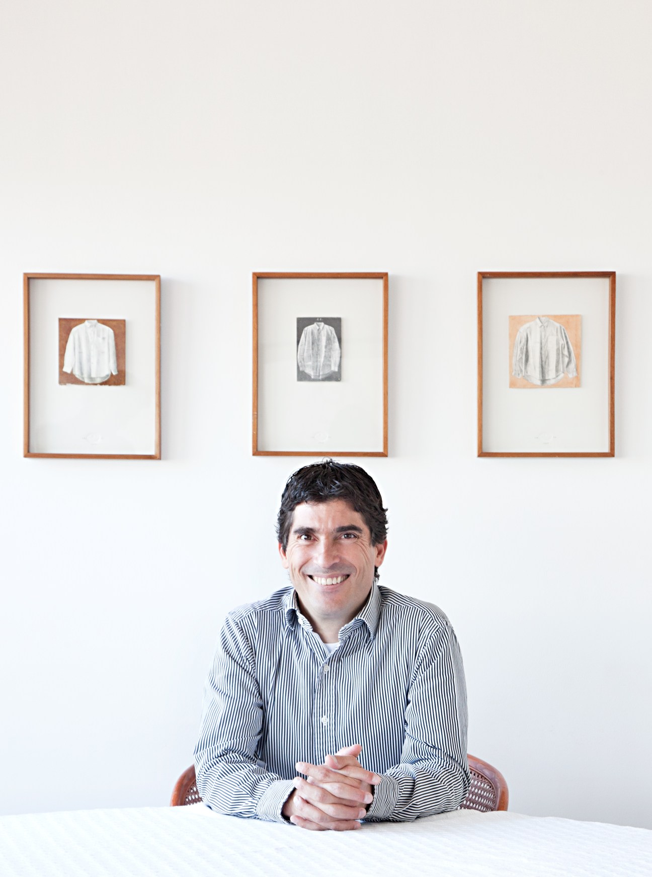 Juan Camilo Sierra, manager of Park Towers cultural centre, seated at a table smiling with framed shirt drawings on the wall behind him.