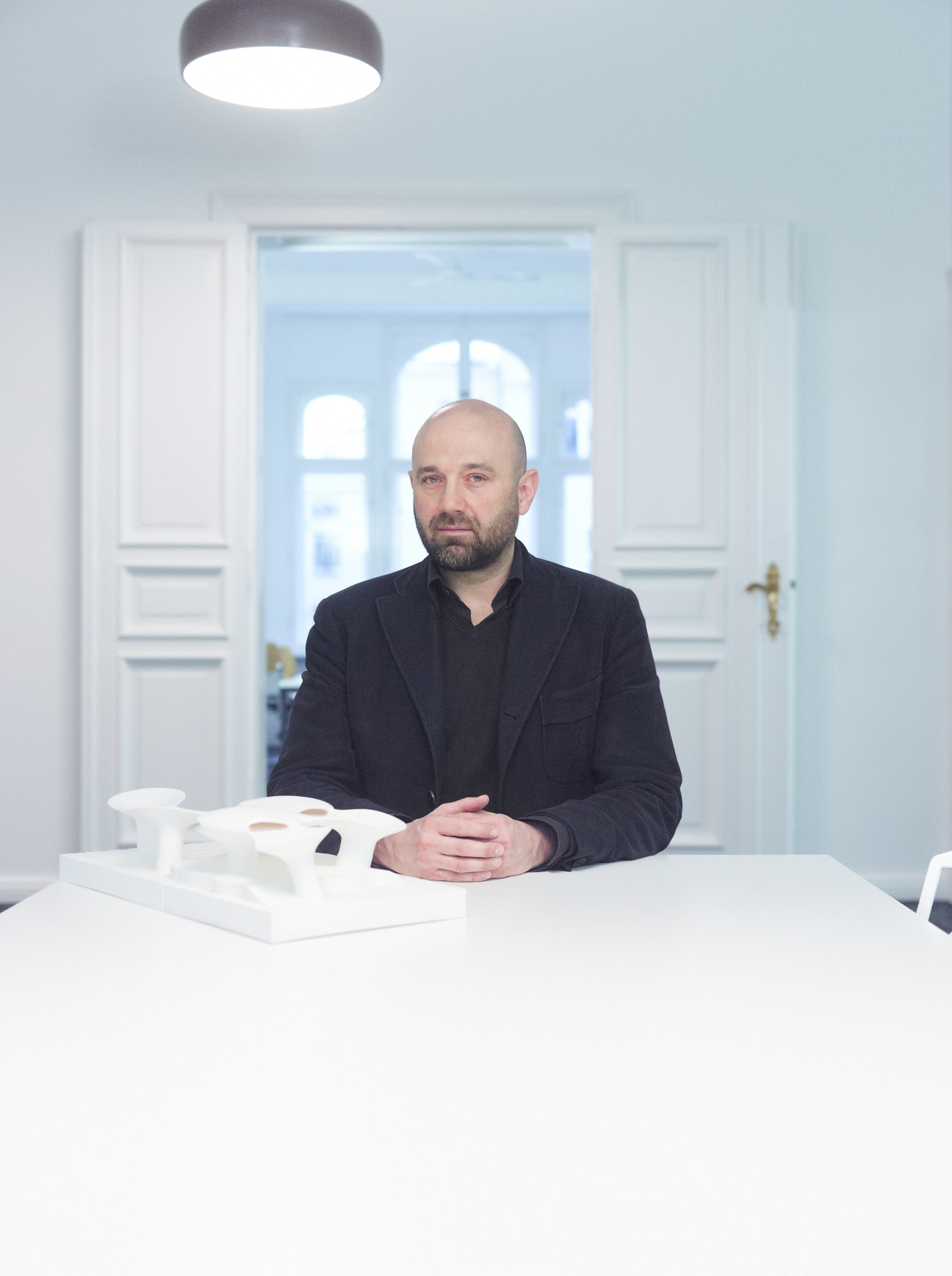 Architect Juergen Mayer H seated at a white table in a minimalist office with white doors and modern ceiling light.