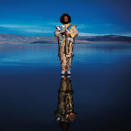 Kamasi Washington holding a saxophone while standing in water against a blue sky backdrop.