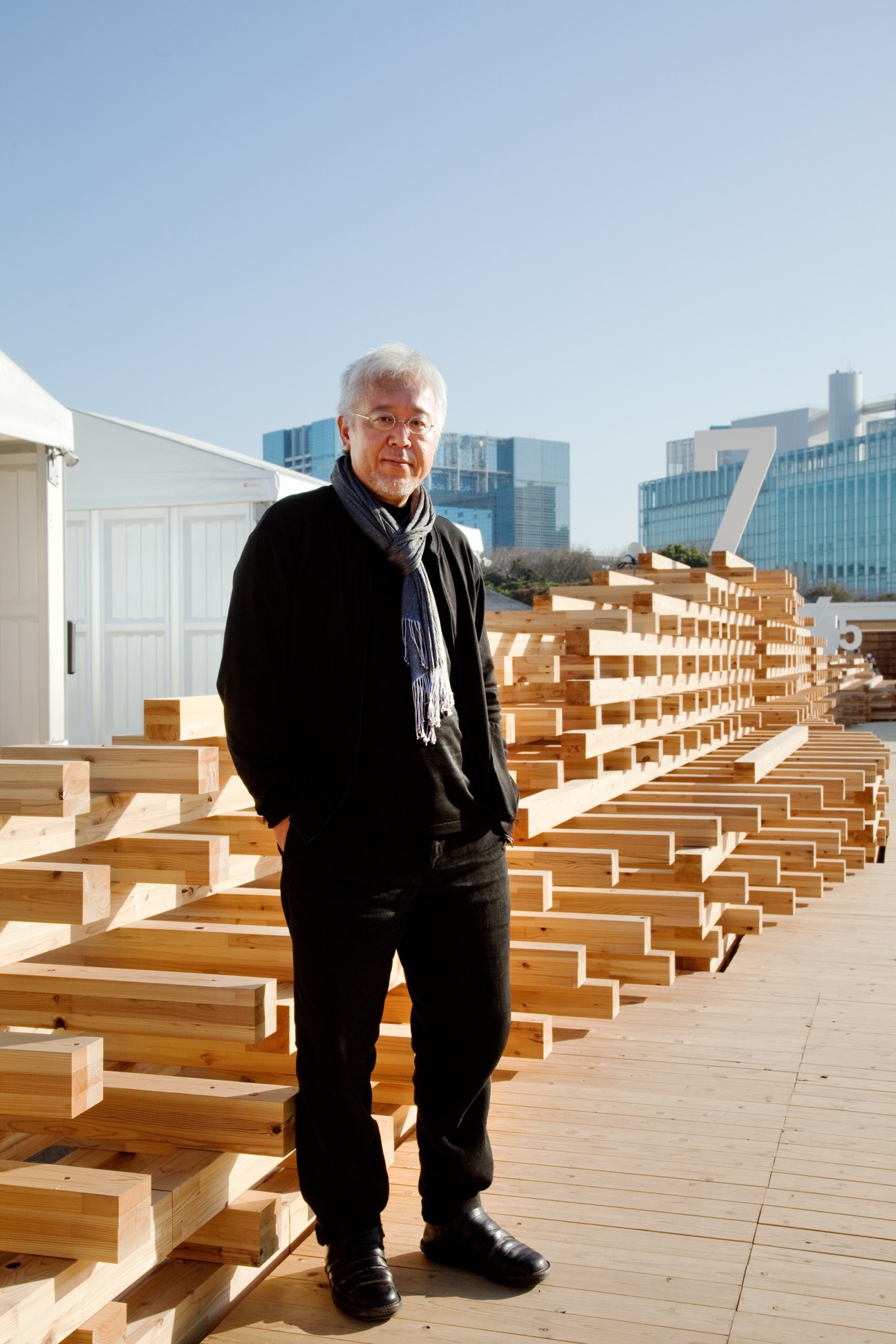 Kenya Hara stands among stacked wooden beams on a rooftop with Tokyo skyline behind him.